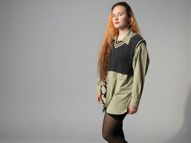 VertieTllo has long, wavy orange hair, green and black dress with lace-up sides, black stockings, and a confident, slightly sideways stance. She's looking to the side.