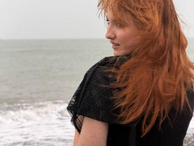 JenniferOhWow has red, wind-blown hair, wearing a black, net-like top, looking over her shoulder with a slight smile, by the ocean. Her ass is partially visible.