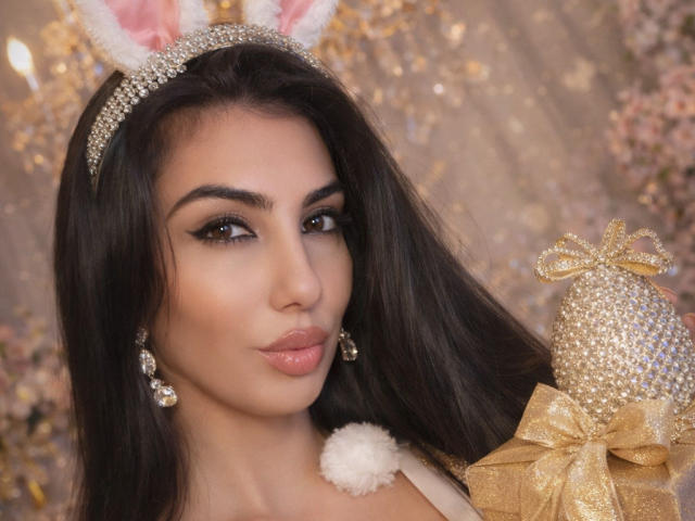 BrunetteBabe6 is a close-up portrait of a woman with long dark hair, wearing a sparkly headband with white, furry bunny ears. She has defined eyebrows, winged eyeliner, and glossy pink lips. She holds a gold, glittery ornament with a bow, in her right hand. She wears pearl earrings and a white, fluffy bunny tail is visible on her chest. The background is blurred with golden sparkles. Her makeup is bold, and her skin tone is warm. The image has a glamorous, festive feel.