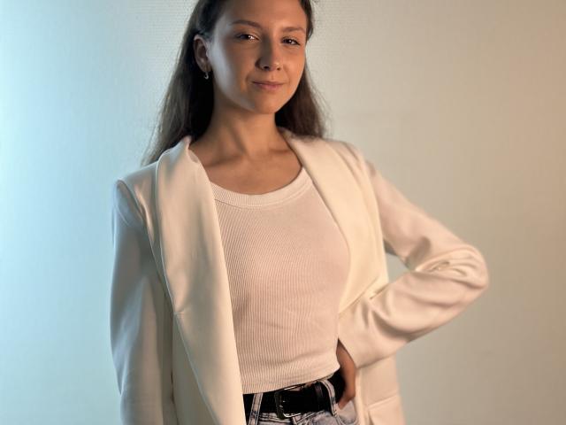 DainaFeldner with long brown hair, white shirt, and open white blazer, standing, hand on hip, subtle smile, light skin, slim, small tits, black belt, neutral background.