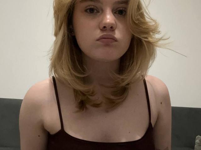 ChristopherSzala with medium-length blonde hair, wearing a brown spaghetti strap top, has a neutral expression, and is showing her tits. Her ass is not visible.