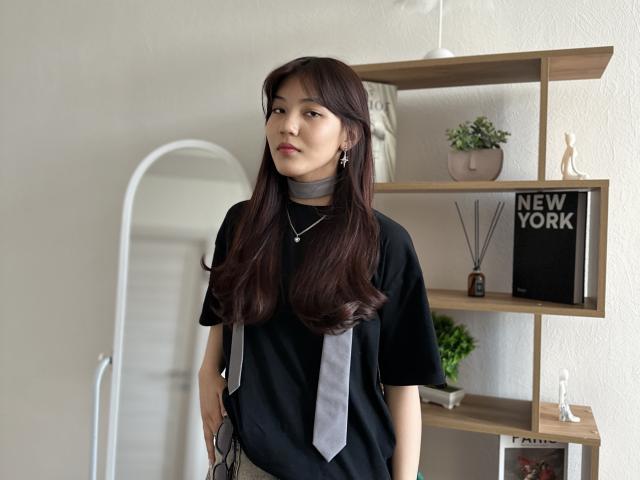 HilariaLedwig with long brown hair, black t-shirt, grey scarf, and necklace, standing in a room with wooden shelves, holding glasses in her right hand.