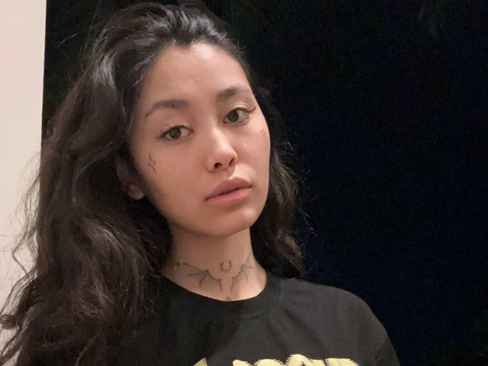 CrisSkalsky with long black hair, light brown skin, and tattoos on the neck and cheek, wearing a black shirt, has a neutral expression, and brown eyes.