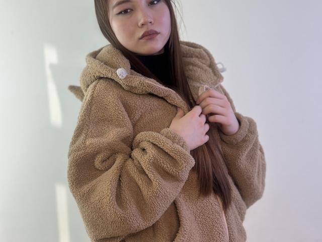 NereidaBottino stands in the frame, wearing a brown, fuzzy, hooded jacket with a small, round, white button on the left side. She has long, straight brown hair, slightly parted to the right, and is looking directly at the camera with a neutral expression. Her right hand is gently touching the jacket's hood. The texture of the jacket is plush and soft, contrasting with her smooth, fair skin. The image has a minimalist, almost clinical, white background. The composition is simple, with NereidaBottino centered and occupying most of the frame.