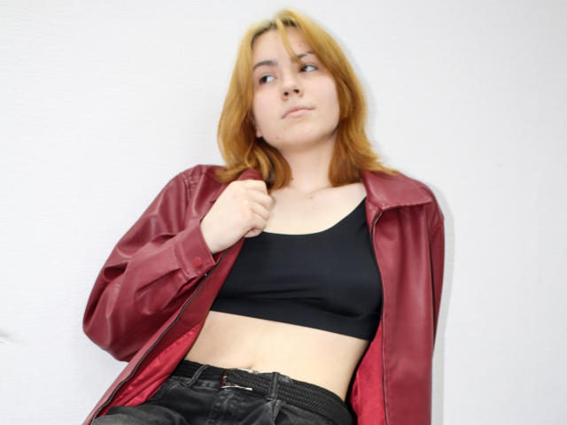 RemyCalhoun with light brown hair, wearing a red shiny jacket, black sports bra, and black jeans, holding jacket collar, looking slightly to the side with a neutral expression.