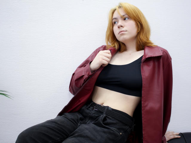 RemyCalhoun with yellow hair, wearing a red jacket, black sports bra, and black pants, sitting, looking to the side, holding jacket, showing a slight belly, relaxed.