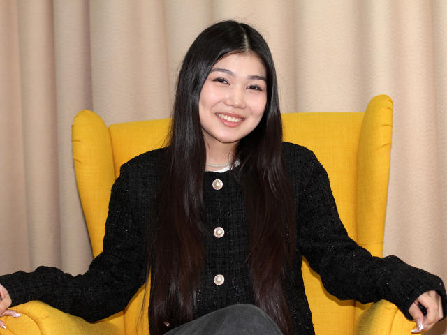 AmeliyaAyrton, Asian woman with long black hair, sits in a yellow armchair. She wears a black knit sweater with white buttons and gray pants. She smiles directly at the camera, her hands resting on the armchair's arms. The image is sharp with warm lighting, highlighting her fair skin and subtle makeup. Her straight hair is parted in the middle.