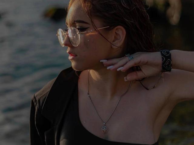 FellDivain with wet brown hair, wearing glasses and a black shirt, touching her neck, showing a necklace with a heart pendant, and a black lace bracelet.