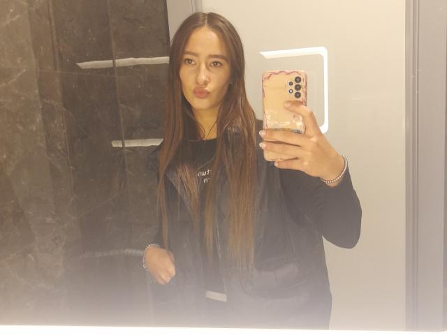 Eine Frau namens Omella mit langen, braunen Haaren, die eine schwarze Jacke und ein schwarzes Hemd trägt, macht ein Selfie mit einem rosa Handy im Badezimmer und zeigt einen neutralen Gesichtsausdruck..