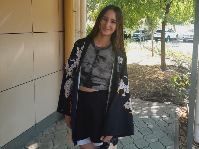 Omella with long brown hair, smiling, wearing a black floral kimono over a grayscale tee, black shorts, and white socks, standing by a yellow wall and hexagonal pavement.