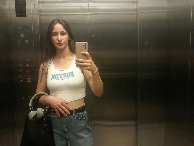 Omella with tits in a white "Motorhead" tank top, blue jeans, holding a phone, and a black bag, standing in a metal elevator, with headphones in her other hand.