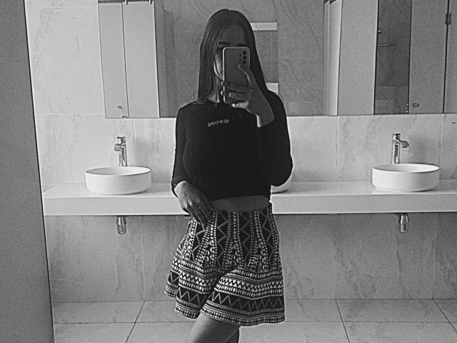 Omella with tits covered by a black long-sleeve top, holding a phone in front of her face, wearing a patterned skirt, standing in a bathroom with sinks.
