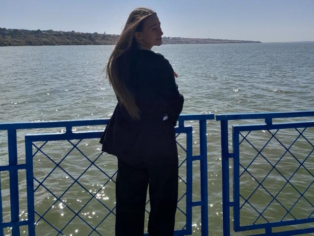 Omella, con lunghi capelli castani, giacca nera e pantaloni neri, è in piedi su una ringhiera di metallo blu, guardando l'acqua verdognola, con il sole che le illumina la schiena, in una giornata tranquilla..
