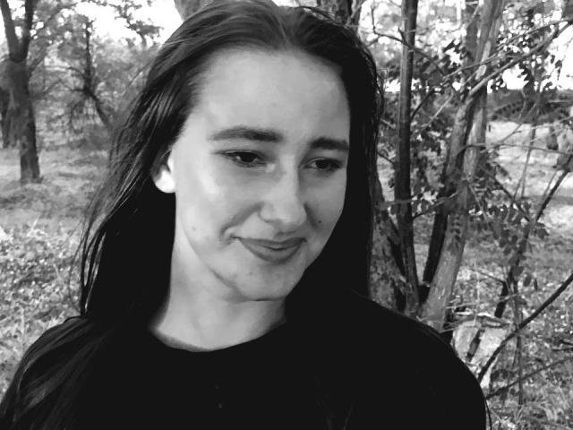 Black-and-white photo of Omella with long, wet, dark hair, slight smile, and small, round ass, wearing a black shirt, standing in a forested area.