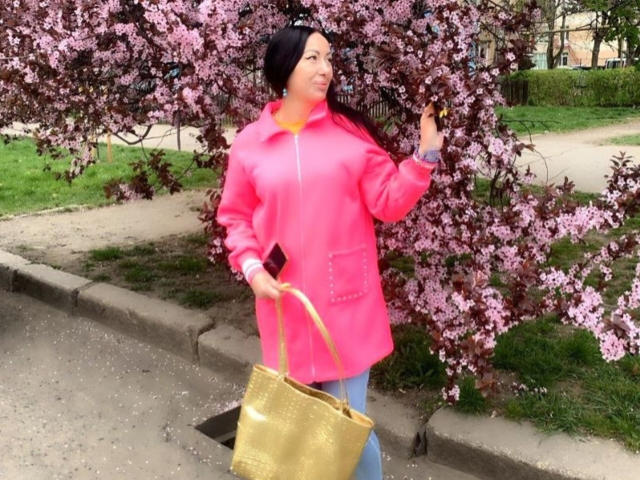 MirandaHott6 stands in front of a cherry blossom tree, wearing a bright pink jacket and blue jeans. She holds a gold shopping bag in her right hand and a black phone in her left. Her dark hair is partially covered by a black headband. The cherry blossoms are pink with dark red leaves. The path she's standing on is gray and slightly uneven. The grass in the background is green.