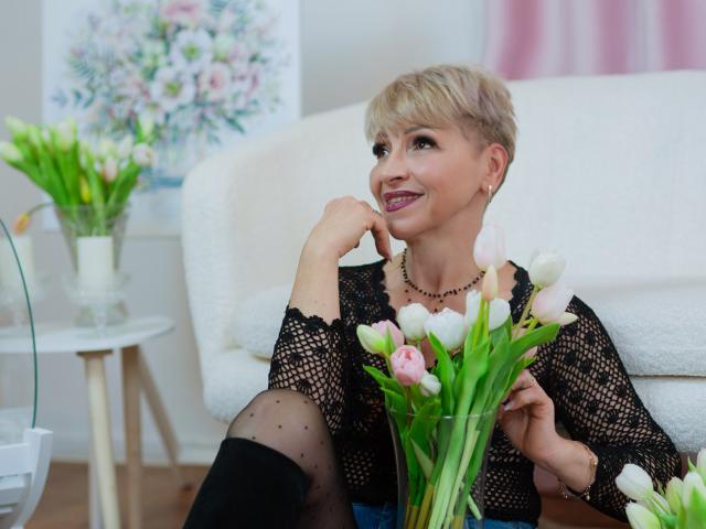 EllaBonito with short blonde hair, black lace top, and polka-dot tights, smiling, holding pink and white tulips, sitting on white couch, right hand on cheek.