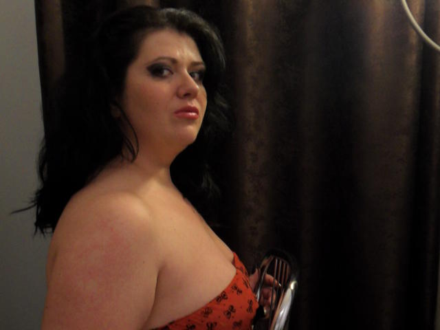 Carolina69 with dark hair, red strapless dress, and bare shoulders, looking sideways with a slight frown. Her tits are partially visible. She has a red mark on her right shoulder.
