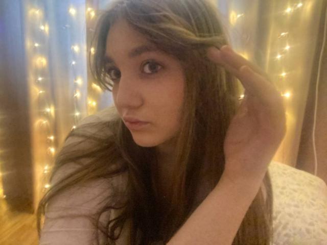 DejaFrayer with long brown hair, big brown eyes, and a serious expression, touching her hair with one hand, wearing a gray top, and surrounded by warm fairy lights.