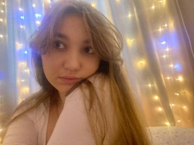 DejaFrayer with long brown hair, slightly wavy, wearing a white shirt, looking at the camera with a subtle smile, soft fairy lights in the background.