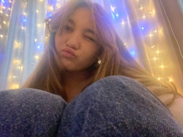 DejaFrayer with long brown hair, winking, and pouting, wearing blue textured pants, sitting with knees up, surrounded by glowing blue and yellow fairy lights.