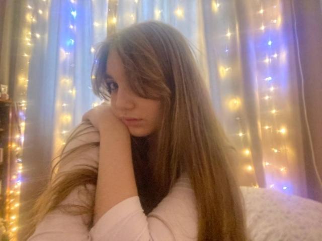 DejaFrayer with long brown hair, resting chin on hand, looking down with a pouty face, wearing a white long-sleeve shirt, surrounded by warm fairy lights.
