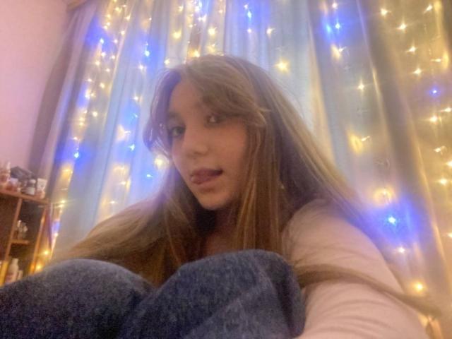 DejaFrayer with long brown hair, blue eyes, and a cute face, tongue slightly out, wearing a white shirt, sitting with knees up, surrounded by glowing fairy lights.
