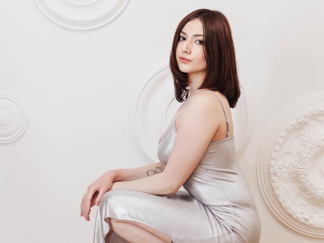 KaylaDirtyLove with shoulder-length brown hair, wearing a shiny silver dress, sitting with her ass slightly raised, looking over her shoulder with a subtle smile. Tattoo on her right arm.