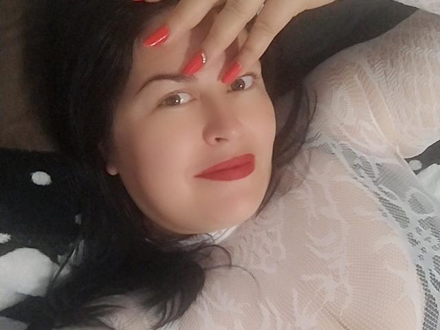 Marryssa69 with red nails, black hair, and red lipstick, wearing a white lace top, lying down, smiling slightly, covering her forehead with her hand. Boobs visible under lace.