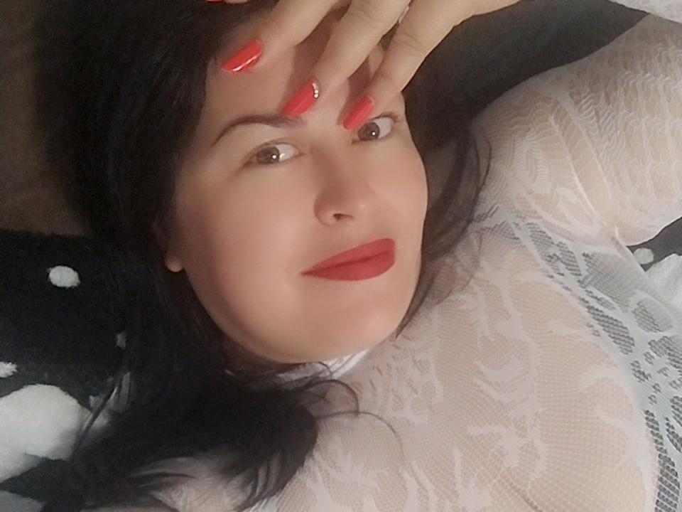 Marryssa69 with red nails, black hair, and red lipstick, wearing a white lace top, lying down, smiling slightly, covering her forehead with her hand. Boobs visible under lace.