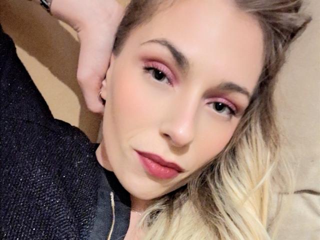 LadyNorth mit rosigem Lidschatten, rotem Lippenstift und blonden Haaren, liegend, trägt ein schwarzes, strukturiertes Oberteil, eine Hand hinter dem Kopf, ein leichtes Lächeln, leicht zerzaustes Haar, weiche Gesichtszüge..