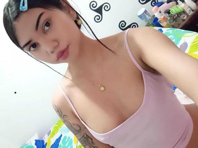 VyxiraMarchan, con pechos pequeños, una camiseta rosa, un tatuaje en el brazo derecho, cabello oscuro sujeto con pinzas, un collar dorado, maquillaje discreto y una expresión seria, en un dormitorio..