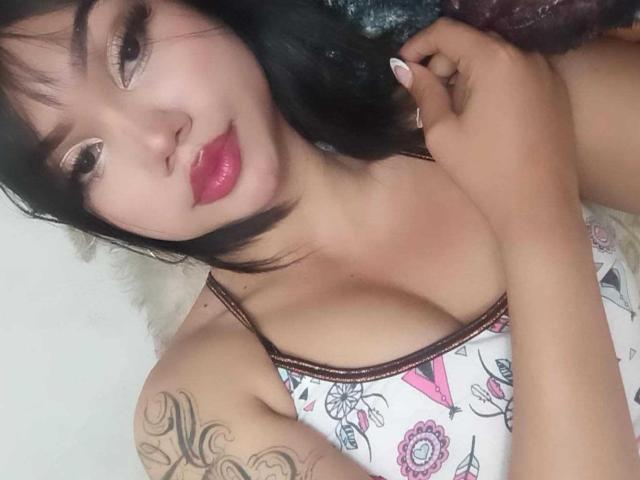 Vyxira, con cabello negro, lápiz labial rojo y un vestido blanco con estampado floral, mostrando tatuajes en el brazo derecho, sujetándose el cabello, con un busto grande y un rostro atractivo, en un selfie..