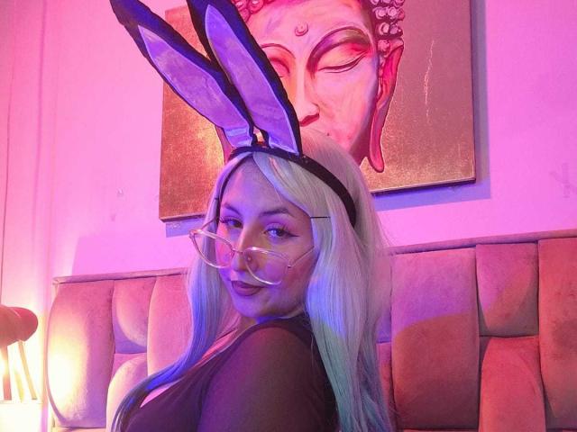 MaddieLesYoux with long blonde hair, wearing black bunny ears and glasses, looking over her shoulder, showing her ass, in a pink and purple lit room.
