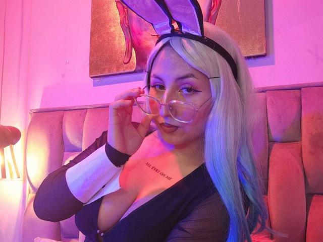 MaddieLesYoux has long, straight, platinum blonde hair and wears a black headband with white bunny ears. She's adjusting oversized, clear-framed glasses with her right hand. Her outfit includes a black, sheer top with white cuffs and a low-cut neckline, revealing a tattoo on her right chest that reads "I am a slave." Her makeup is subtle with dark eyeliner and pink lipstick. The pink and purple lighting casts a soft glow on her face and the beige, tufted headboard behind her. The image has a playful, cosplay vibe.