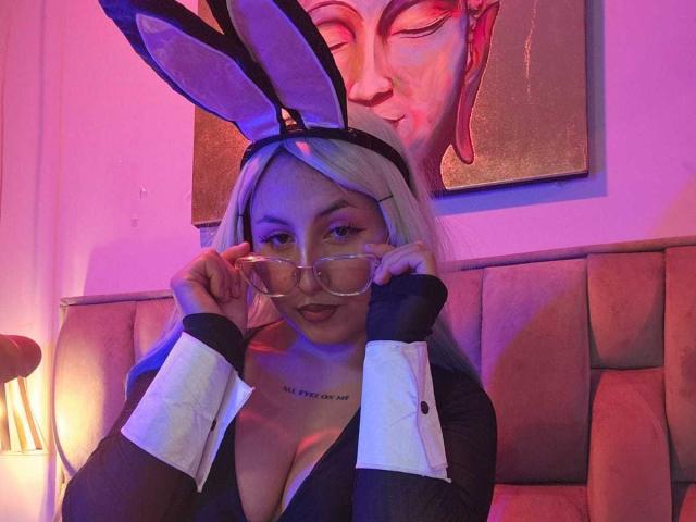 MaddieLesYoux with big tits, wearing a black bunny outfit, holding glasses, purple and pink lights, white cuffs, and black bunny ears, sitting in a cozy, lit-up booth.