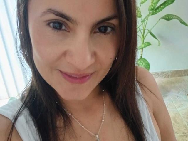 Close-up of SolaraDubois, a woman with light skin, straight dark brown hair, and blue eyes. She has a small silver nose ring in her right nostril and is wearing a white sleeveless top with a delicate silver necklace. Her pink lipstick contrasts with her natural look. Green leafy plant in the background. Textured beige tile floor. Bright light from the left. SolaraDubois is smiling slightly.