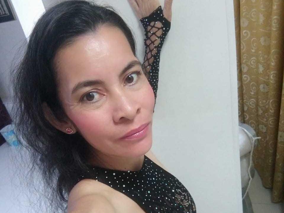 VictoriaGrey with dark hair, pink blush, shiny pink lips, wearing black lace- patterned top, arms raised, standing against white wall, small pink earrings, subtle smile, slightly flushed face.