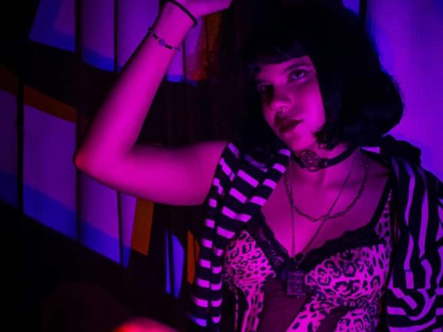 RavenMorninsgstar, con cabello negro y rizado, camisa a rayas, sujetador de estampado de leopardo, collar y colgante, levantando el brazo bajo luces de neón moradas y azules, luciendo seductora. Se ven los senos..