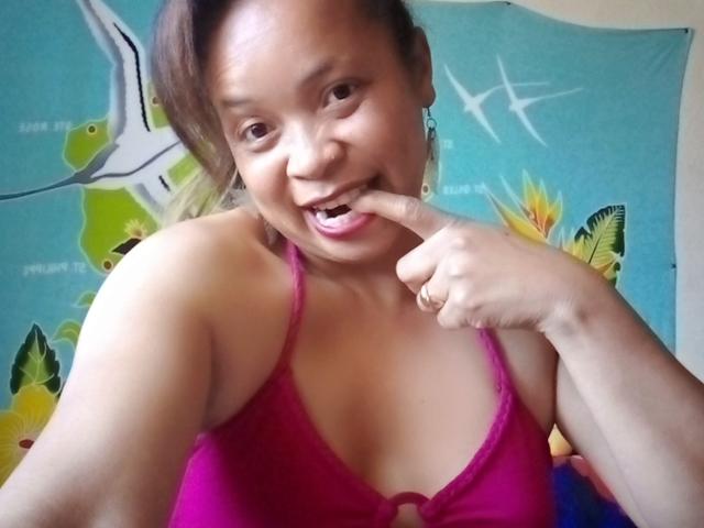 Rahel69 with medium brown skin, smiling, has finger in mouth, wearing pink bra, showing her tits, short brown hair, earrings, and cute face, in a selfie.