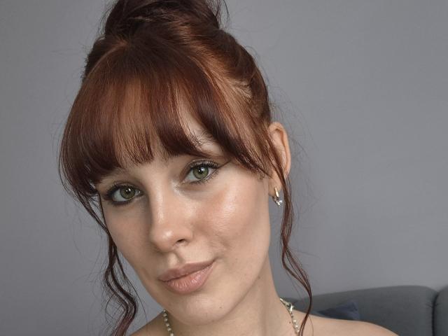 CleoErgaX has red auburn hair in a messy bun, green eyes, subtle makeup, and a slight smile. She's wearing small earrings and a pearl necklace.