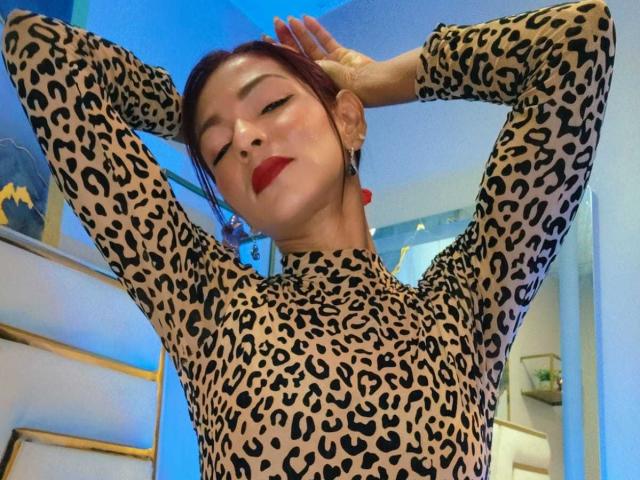 HannaPretty stands with arms raised, hands behind head, wearing a tight leopard-print long-sleeve top. She has red lipstick, red earrings, and her dark red hair is tied back. Her eyes are closed, and she has a slight, confident smile. The top's pattern is bold with black spots on a beige background. The lighting is blue, adding a cool tone to the image. The composition focuses on HannaPretty's upper body and face. The image has a modern, stylish feel.