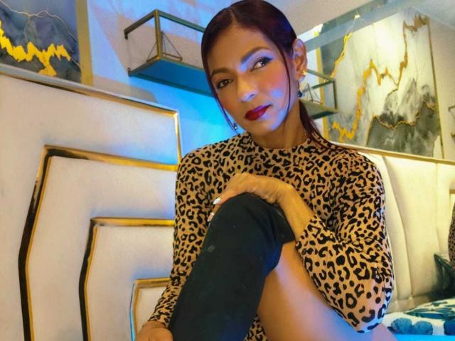 HannaPretty with medium brown skin, straight dark brown hair in a low ponytail, and bright red lipstick. She's sitting, legs crossed, wearing a leopard print long-sleeve top and blue knee-high socks. She's looking slightly to the right with a subtle smile. Background includes a white, gold-trimmed wall with abstract art and shelves. Warm, blue, and yellow light adds depth to the scene. The image is a photograph.