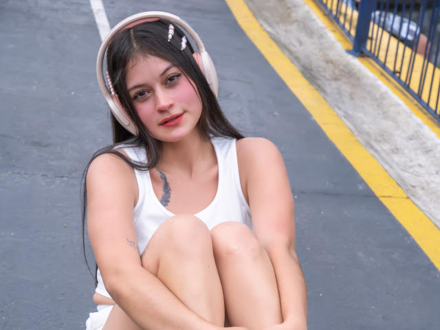 LizzaRose with long dark hair, pink headphones, white tank top, and bare legs, sitting on a road, knees up, arms around legs, slight smile, small tattoos on her arm.