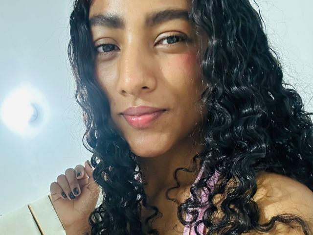 NatalyBlonder with curly black hair, brown skin, and pink lips, holding her shoulder with black-painted nails, wearing a pink top. She's looking at the camera with a slight smile.