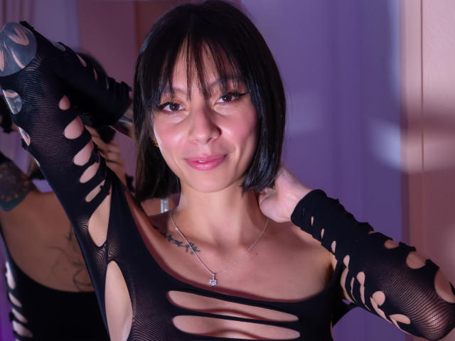 Sara Lambert, con cabello negro corto, vestida con una camiseta negra rasgada que deja ver sus pechos y tatuajes, sonriendo con las manos detrás de la cabeza, llevando un collar, en una habitación iluminada con luz púrpura..