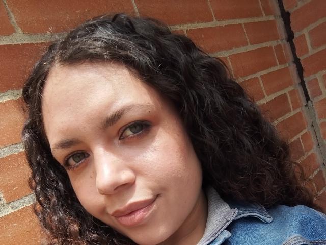 Close-up photo of RubbyStrongX with curly black hair, light brown skin, and green eyes. She has a small mole on her right cheek and is wearing a denim jacket over a gray shirt. RubbyStrongX's eyebrows are thick and slightly arched. She has a subtle, confident smile. The texture of her hair is curly and slightly wavy. The brick wall in the background is reddish-brown with visible mortar lines. The image is sharply focused on her face, with soft lighting highlighting her facial features.