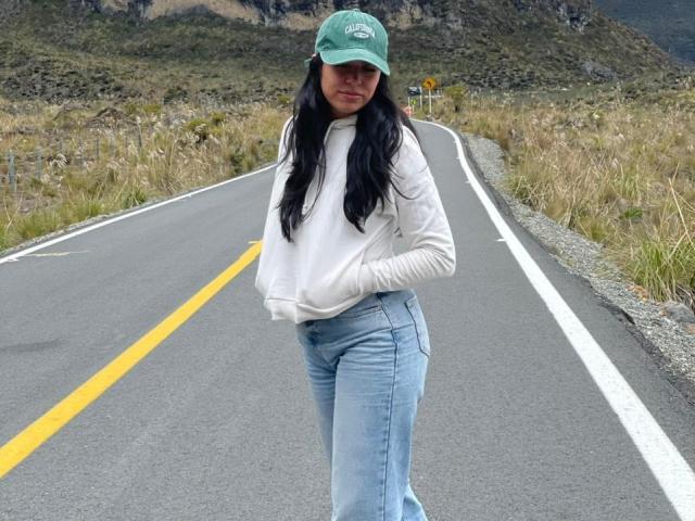 MiluSky stands on a road, wearing a green cap, white hoodie, and blue jeans, with her hands in her hoodie, long black hair, and a slight smile.