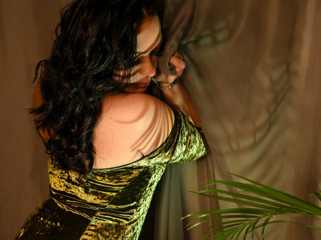 MiaMillerr69 with wavy black hair, green velvet dress, leaning against curtain, shadow on shoulder, green plant in foreground, gold ring on finger, eyes closed, soft lighting.