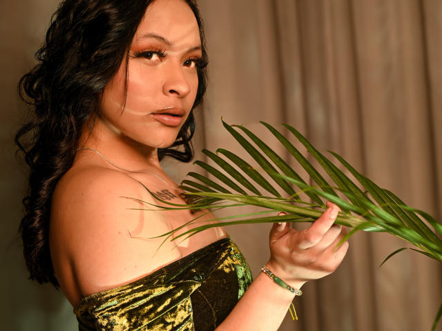 MiaMillerr69 with wavy black hair, holding a green palm leaf, wearing an off-shoulder green velvet top, looking at camera, tan skin, subtle makeup, bracelet on right wrist.