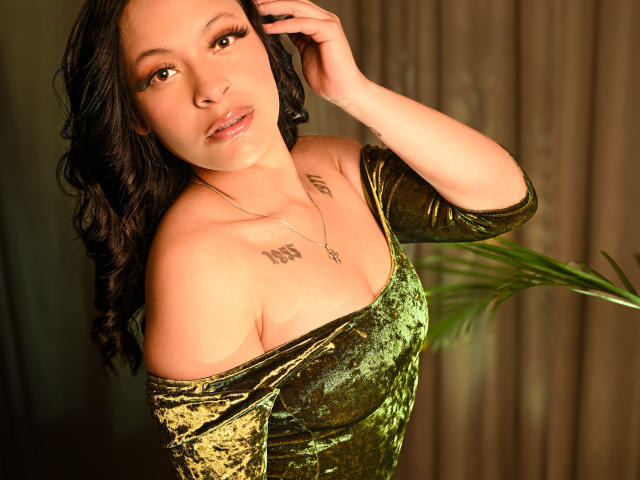 MiaMillerr69 in a green velvet dress, showing off her tits with one arm up, wavy black hair, brown eyes, and a tattoo on her chest. Warm indoor light.